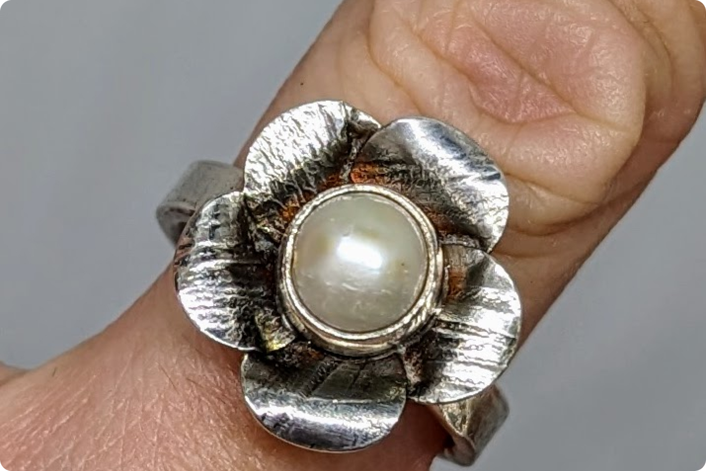 A silver ring shaped like a flower with textured petals and a central round pearl worn on a finger. The background is a plain, neutral tone.