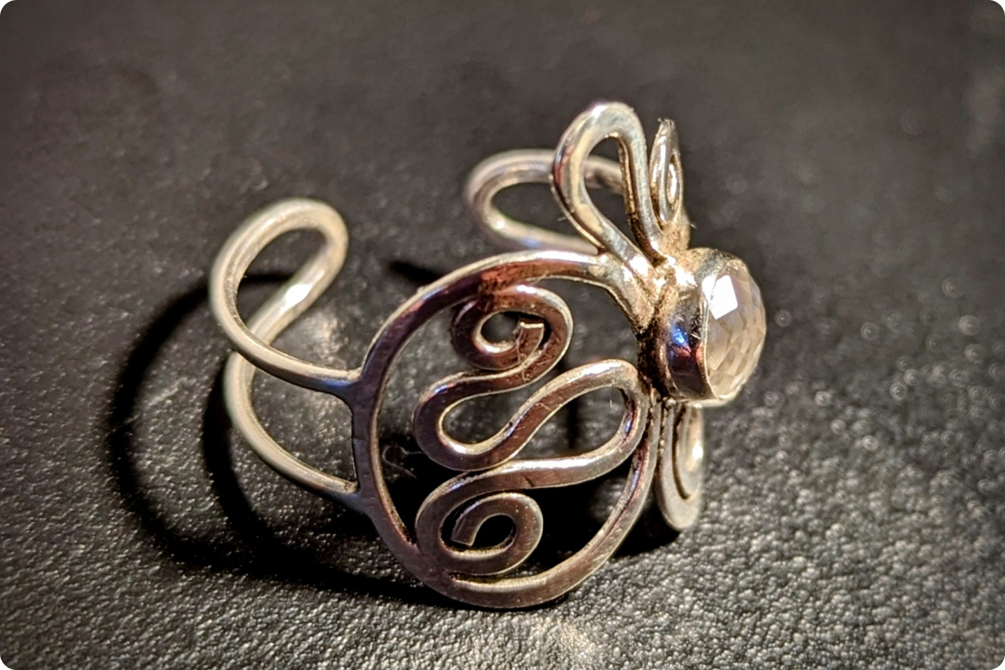 A close-up shot of an ornate silver ring with intricate swirls and a clear gemstone set in the centre. The design features two loops extending on either side of the ring. The background is a dark, slightly textured surface.