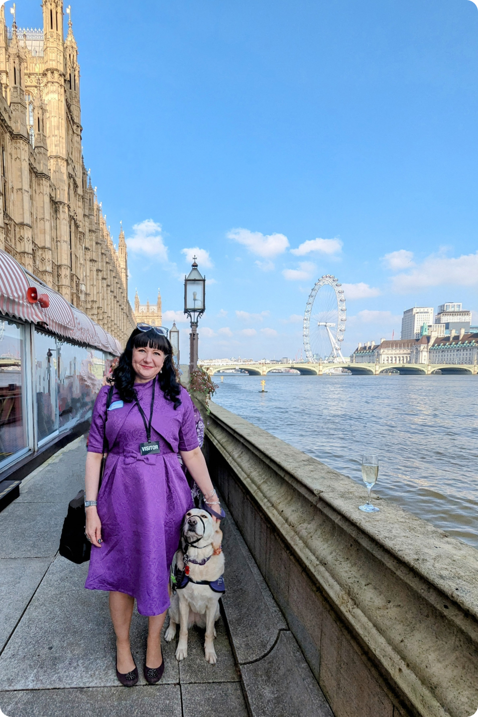 Carol in a purple dress stands next to her assistance dog Bella, on a walkway along the River Thames. The iconic London Eye Ferris wheel is visible in the background, behind the Houses of Parliament. The sky is blue with scattered clouds, and a glass of champagne sits on the ledge.