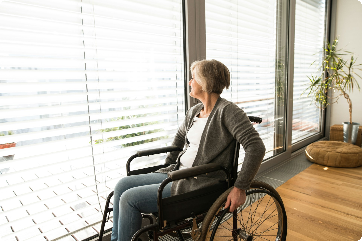 A lady in a wheelchair is gazing out of the window. She has short grey hair and is wearing a mink coloured cardigan over a white top, she is also wearing pale blue jeans.