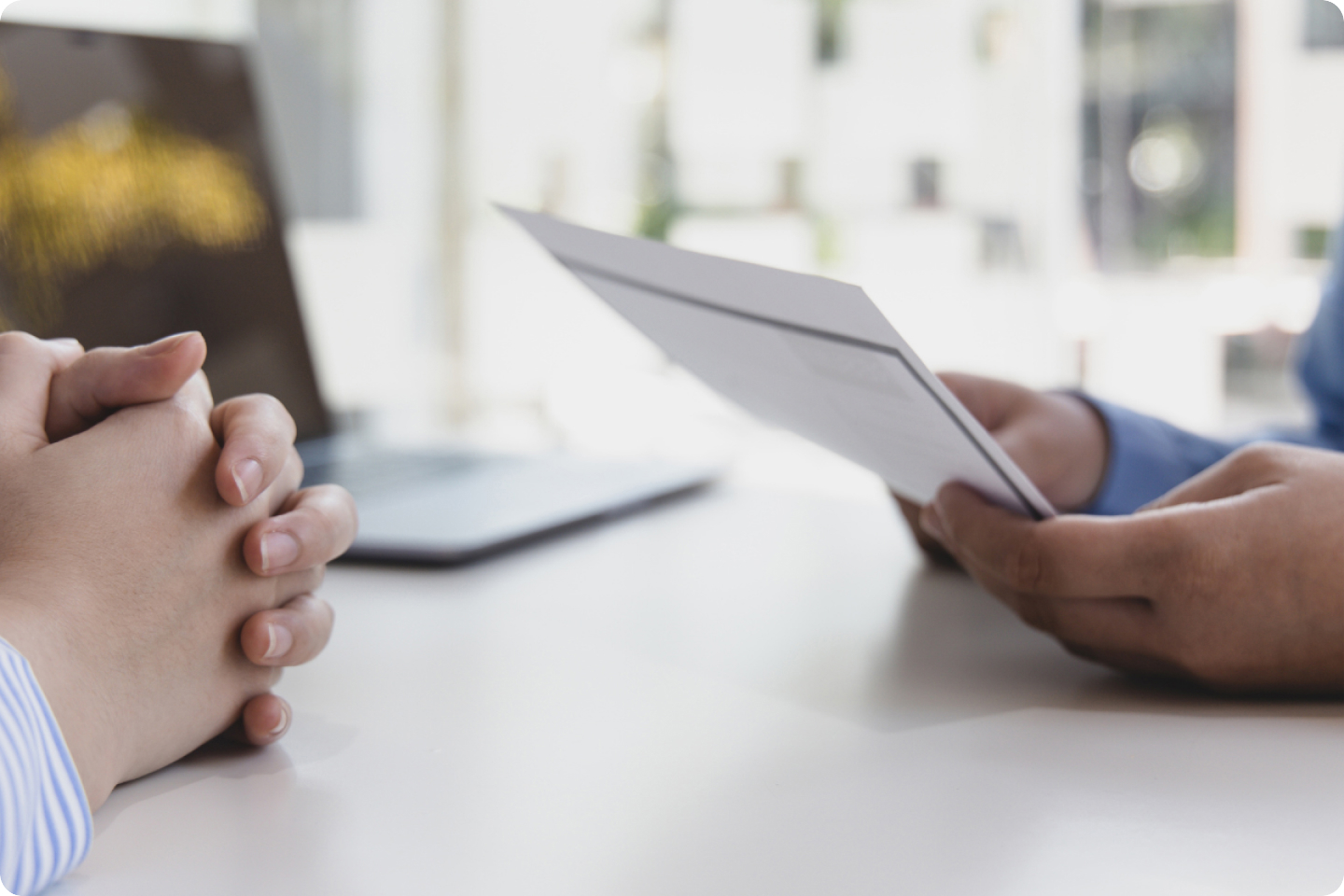 Two people are seated at a table in a well-lit room. One person holds a pair of documents, while the other person has their hands clasped together on the table. A laptop is open in the background. The image suggests a business meeting or interview setting.