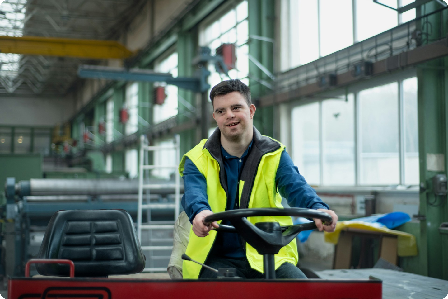 A man wearing a yellow safety vest is smiling while driving a forklift inside a spacious industrial warehouse. The warehouse has large windows and various machinery in the background.