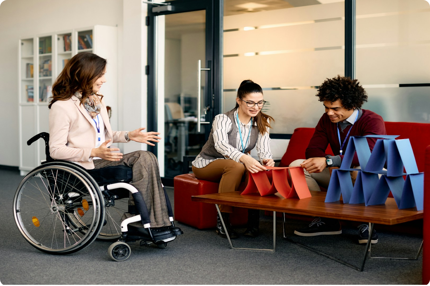 Three people are in an office. A woman in a wheelchair is smiling while two others, a woman with a ponytail and a man with curly hair, build a structure with red and blue paper on a coffee table. They are all wearing lanyards and dressed in business casual attire.