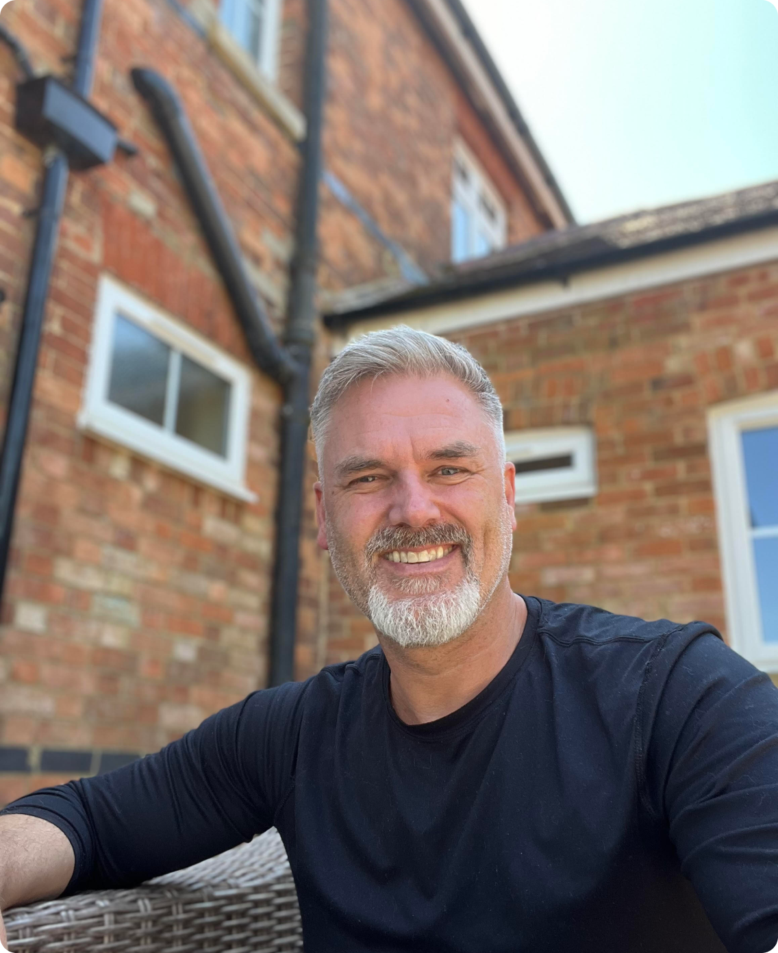 Photo of Gavin smiling at the camera, he has grey hair and beard. He is wearing a navy top and looks very happy. He is standing outside with a brick house behind him.