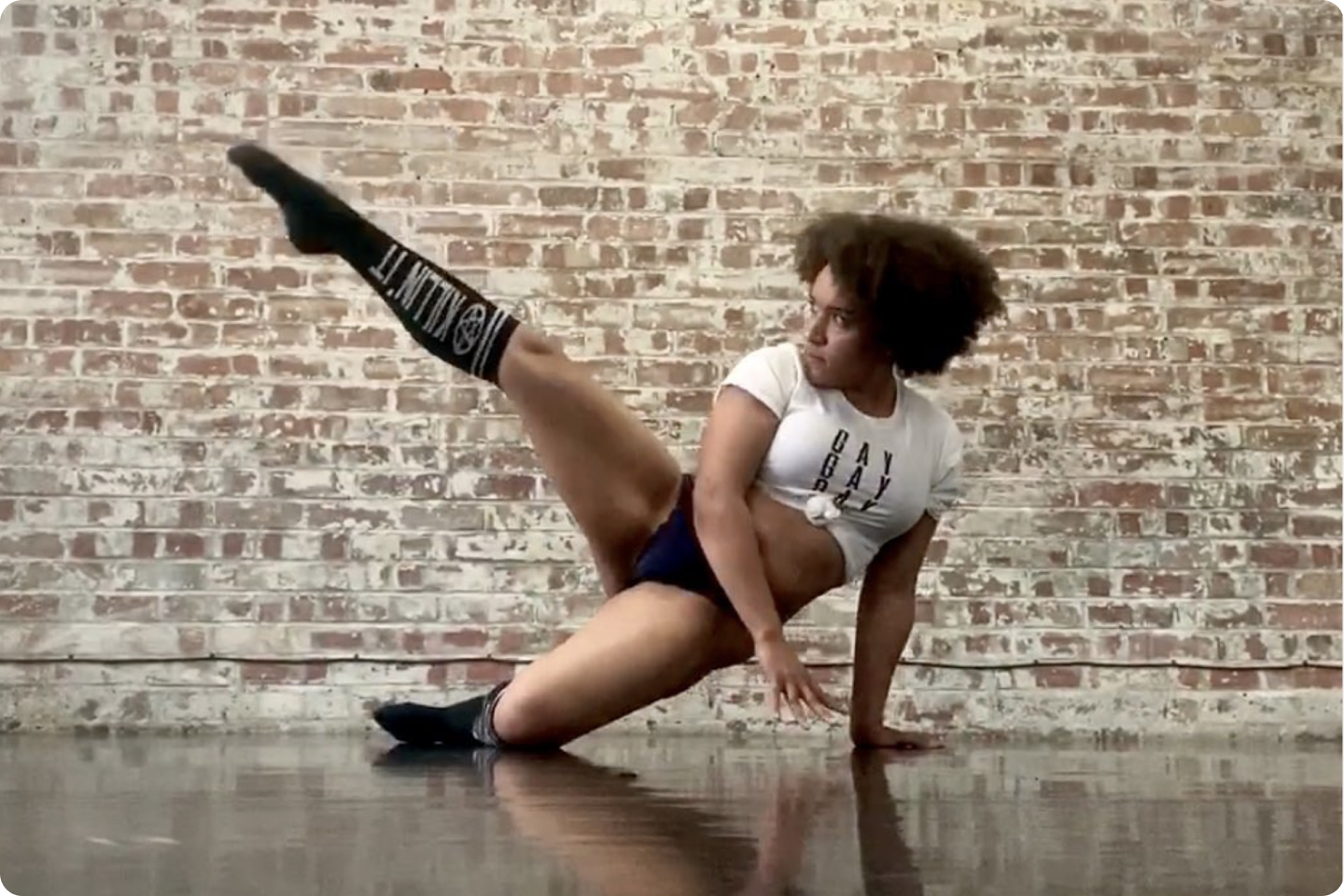 Photo of Georgia in a dynamic pose with one hand pressed against the floor and a leg in the air. Georgia wears a top that says ‘gay gay gay’ and black knee high socks. There is a brick wall in the background.