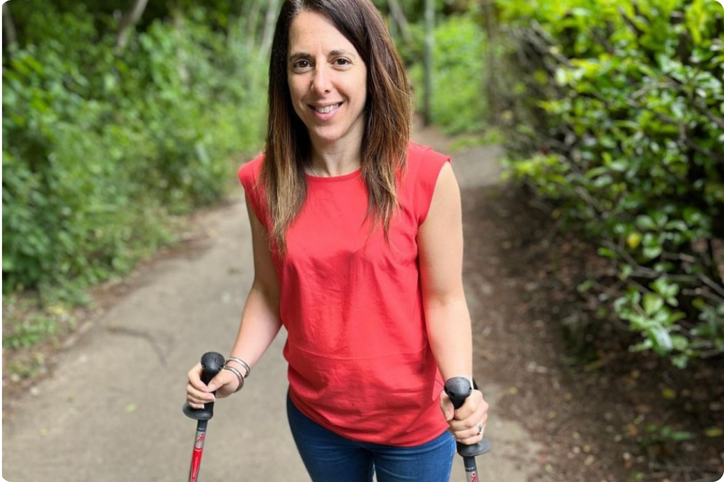 Georgina is a white woman with long dark straight hair, she is wearing a sleeveless red top and jeans and is walking outdoors on a path with green trees and bushes round her and is using two walking sticks