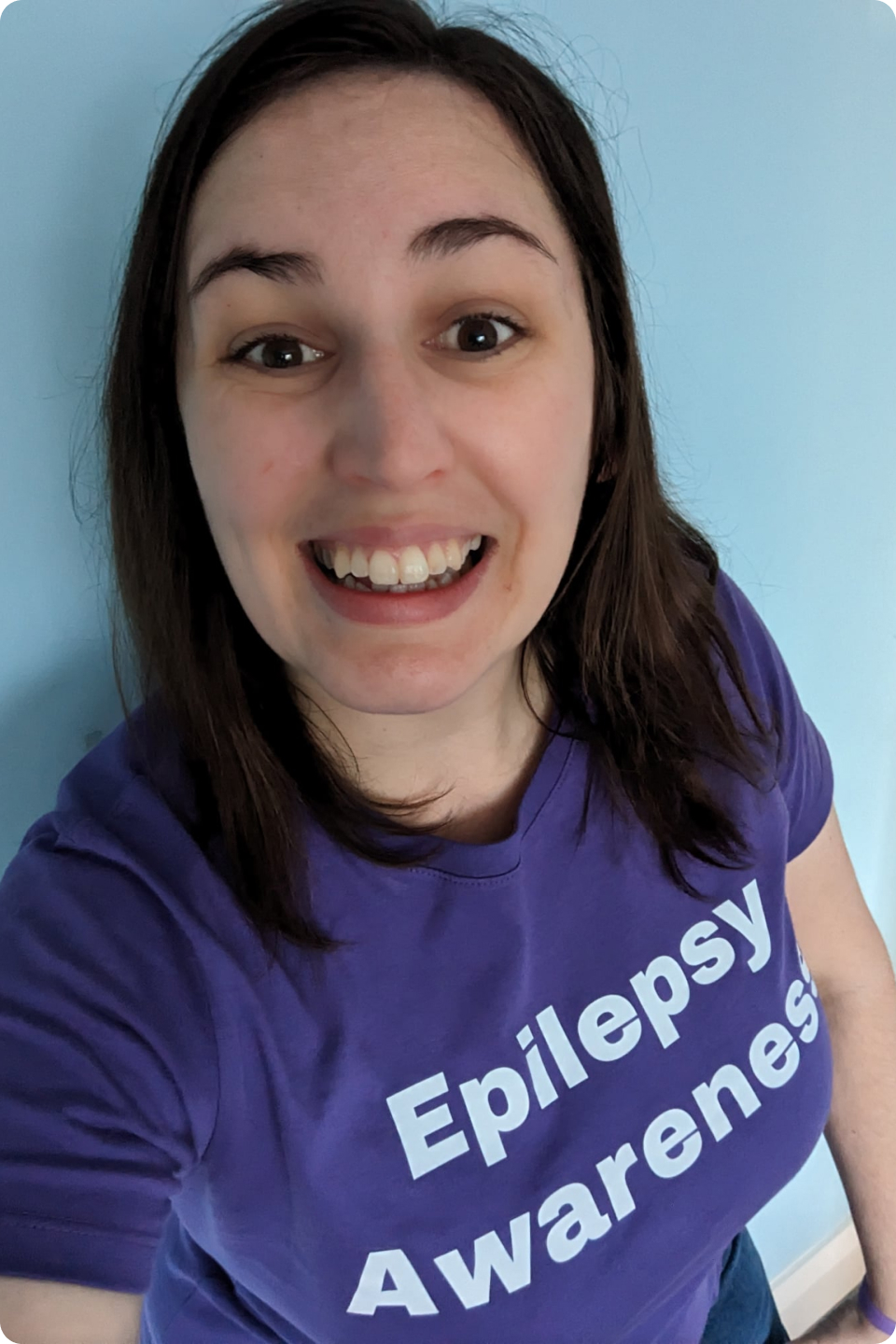 Nicole who has long brown hair is smiling at the camera. They are wearing a purple shirt with "Epilepsy Awareness" written in white letters. The background is light blue