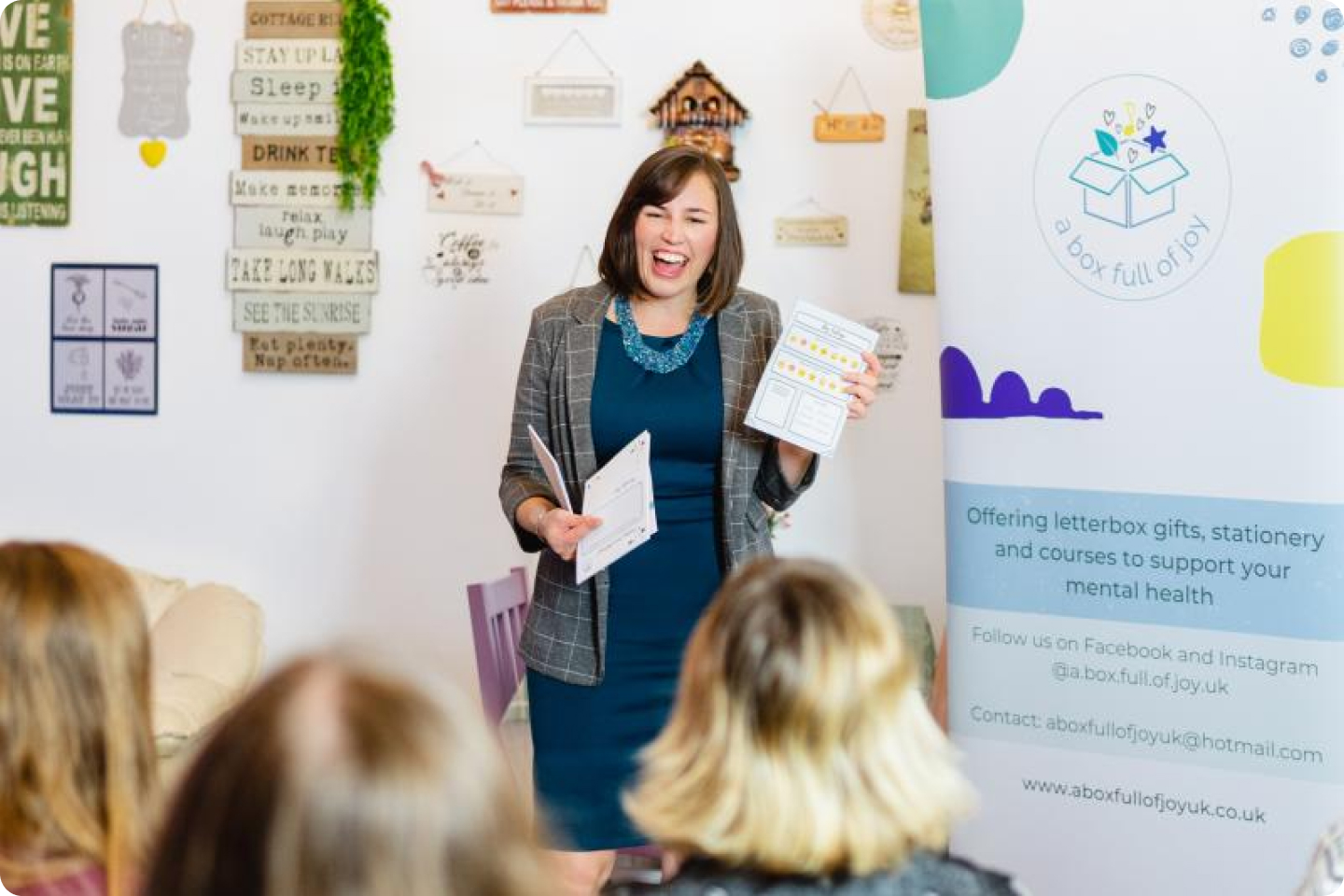 Nicole is wearing a suit jacket over a blue dress whilst speaking to an audience about  ‘A Box Full of Joy’. She is holding some materials in her hand which she is showing the audience. Behind her, there's a banner that reads, "Offering letterbox gifts, stationery and courses to support your mental health." Multiple plaques decorate the wall.