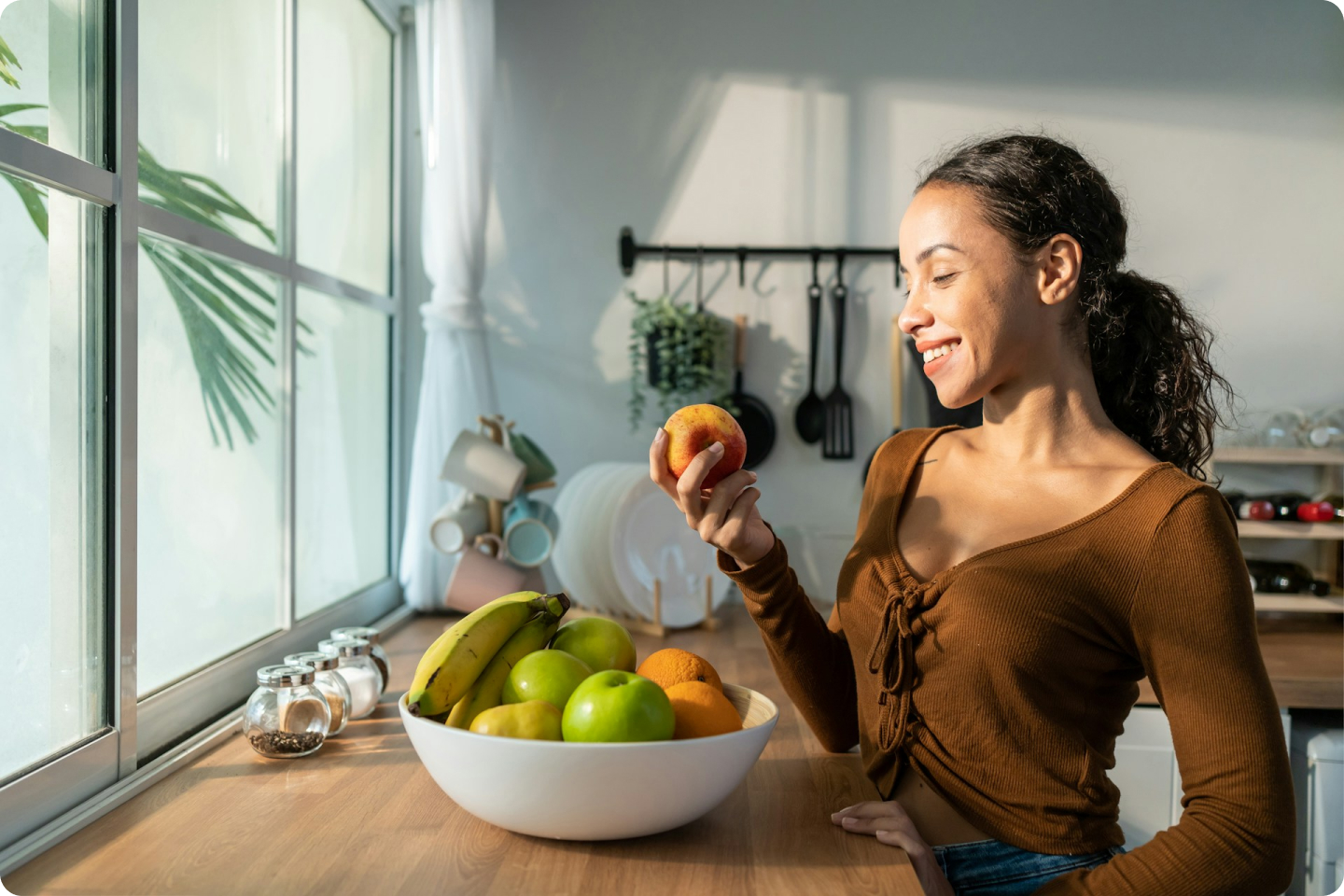 A woman stands in a kitchen, smiling and holding a peach. She is looking out a window. In front of her is a bowl filled with bananas, apples, and oranges. Kitchen utensils and dishes are visible in the background. Natural light illuminates the scene.