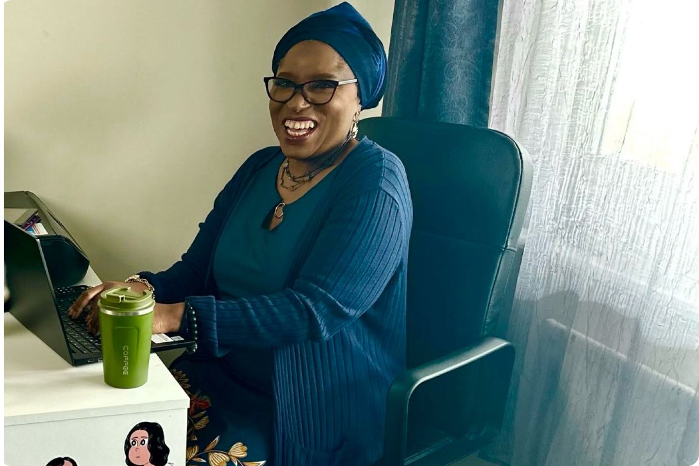 Funmi wearing glasses, a blue headscarf, and a blue cardigan is sitting at a desk with a laptop, smiling at the camera.