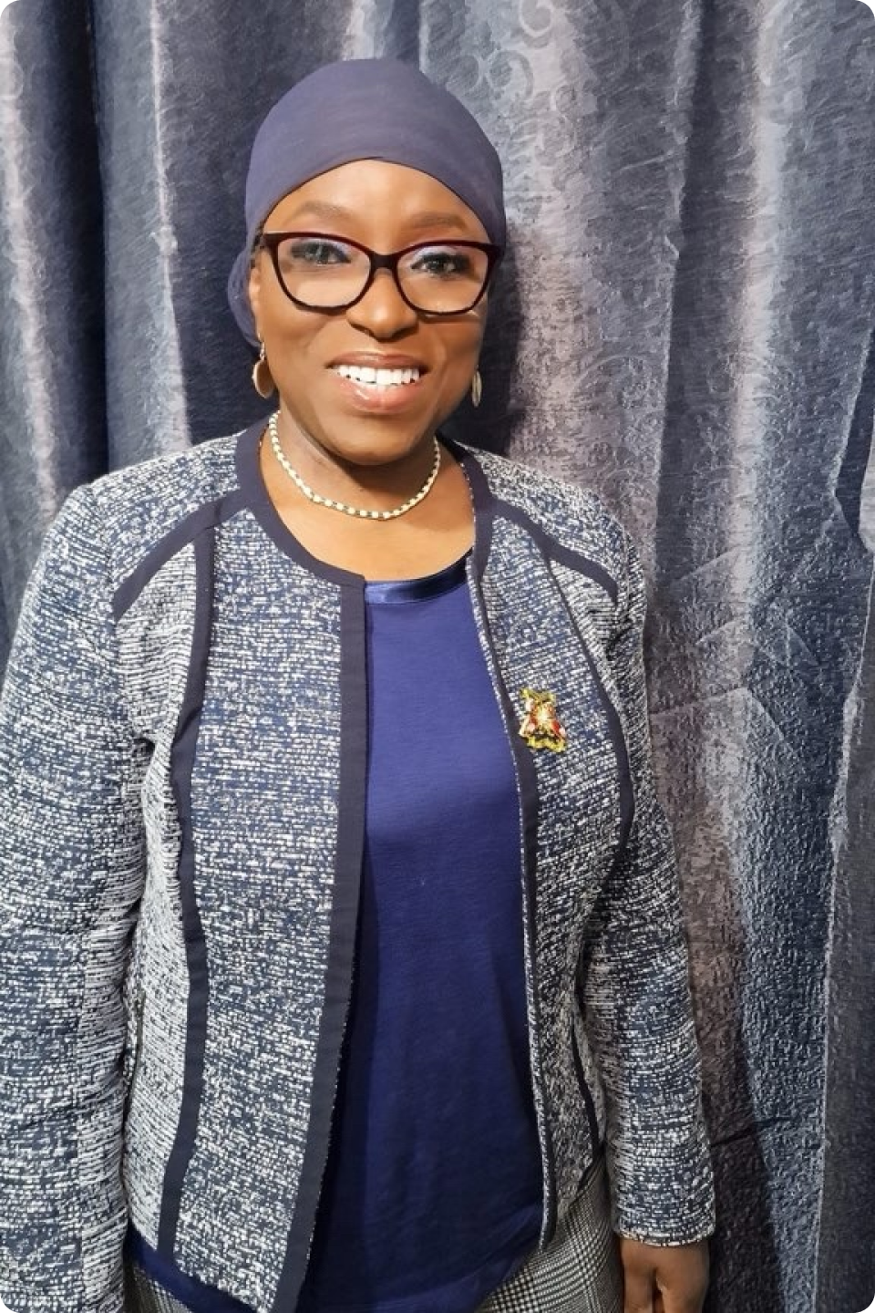 Funmi is wearing a blue headscarf, glasses, a pearl necklace, a blue and grey jacket, and a blue top is standing and smiling in front of a textured blue curtain.