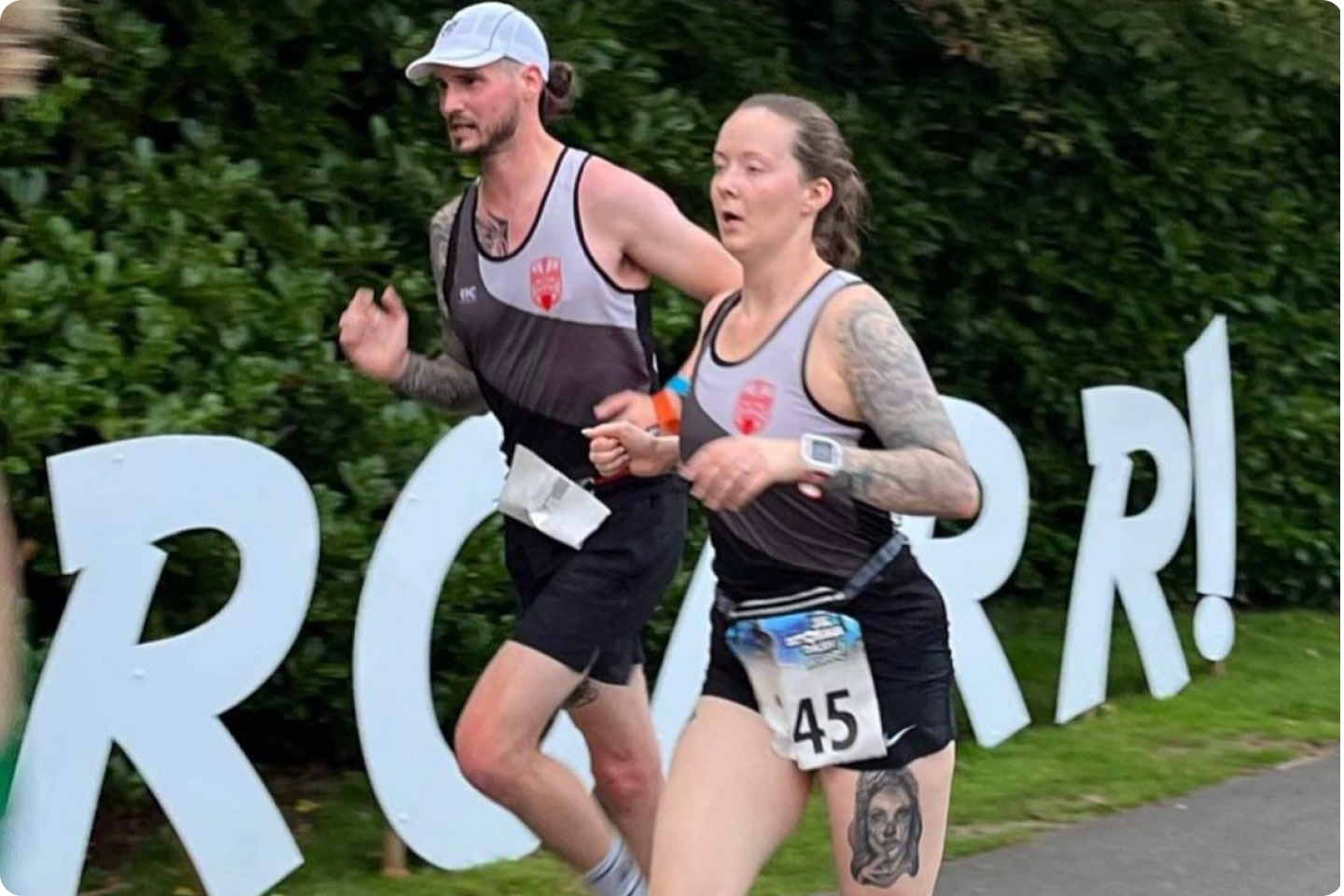 Two runners, a man and a woman, are participating in a race. They both wear black athletic gear with matching logos and have bib numbers, with the woman wearing number 45. The backdrop includes large letters spelling "ROAR!" and a dense green hedge.