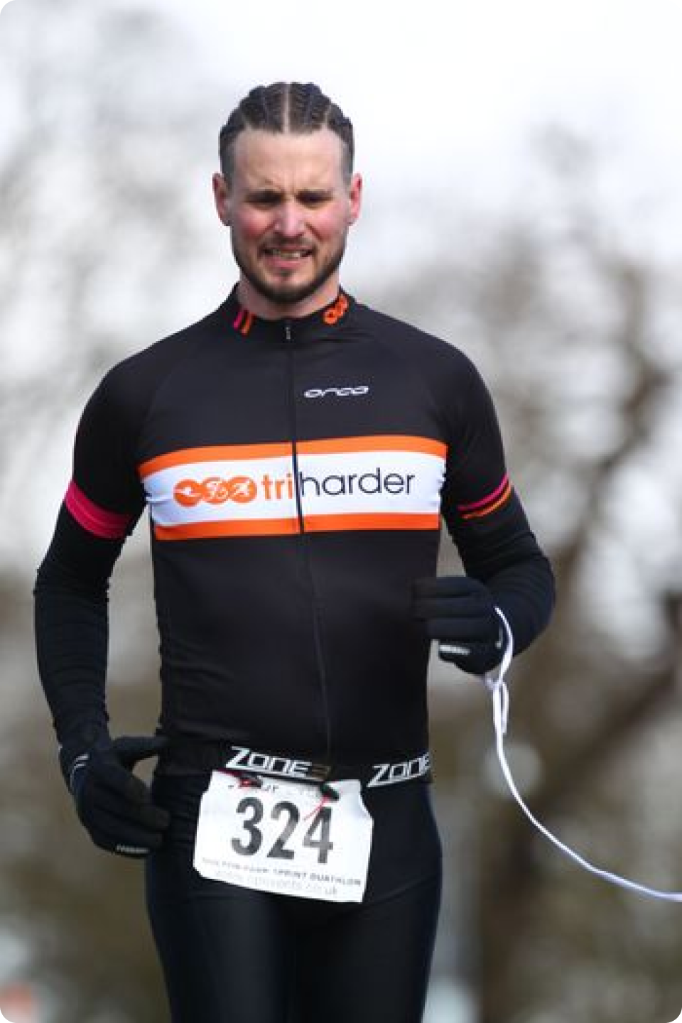 A man is running outdoors. He wears a black athletic jacket with an orange, white, and red stripe and the words "triharder" on it. He has a race bib numbered 324 pinned to his clothing. The background is blurry, featuring bare trees.