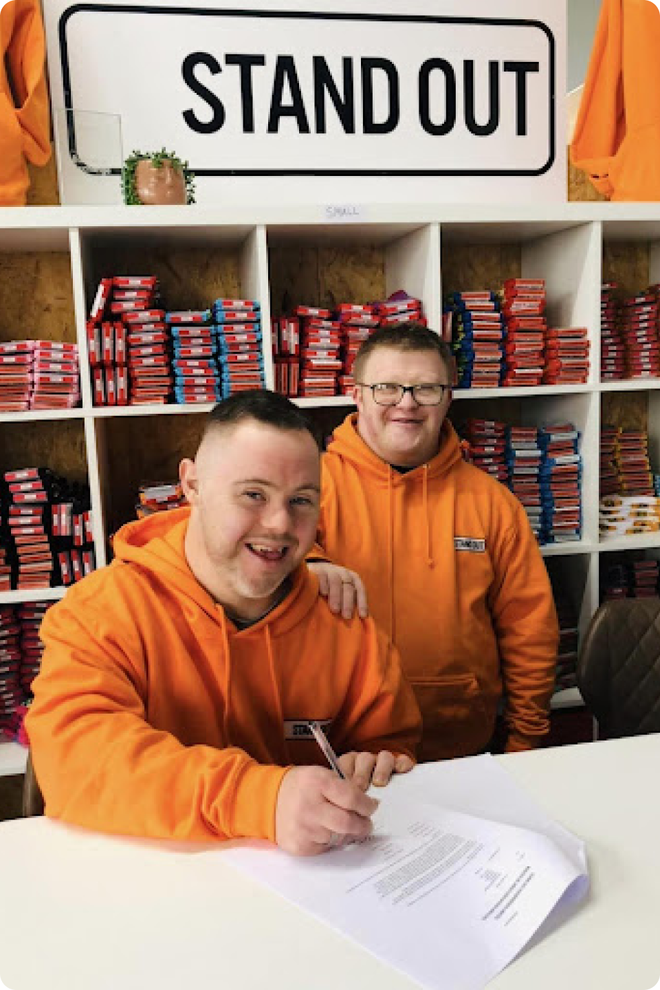 Two men in orange hoodies sitting at a table signing a paper