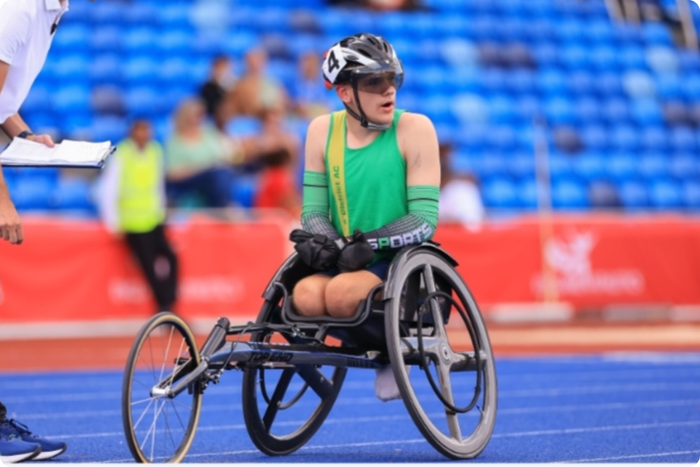 Toby Richardson in a racing wheelchair, wearing a green shirt and helmet, on a blue track with spectators in the background. An official stands nearby holding a clipboard. The athlete appears to be preparing for or finishing a race.