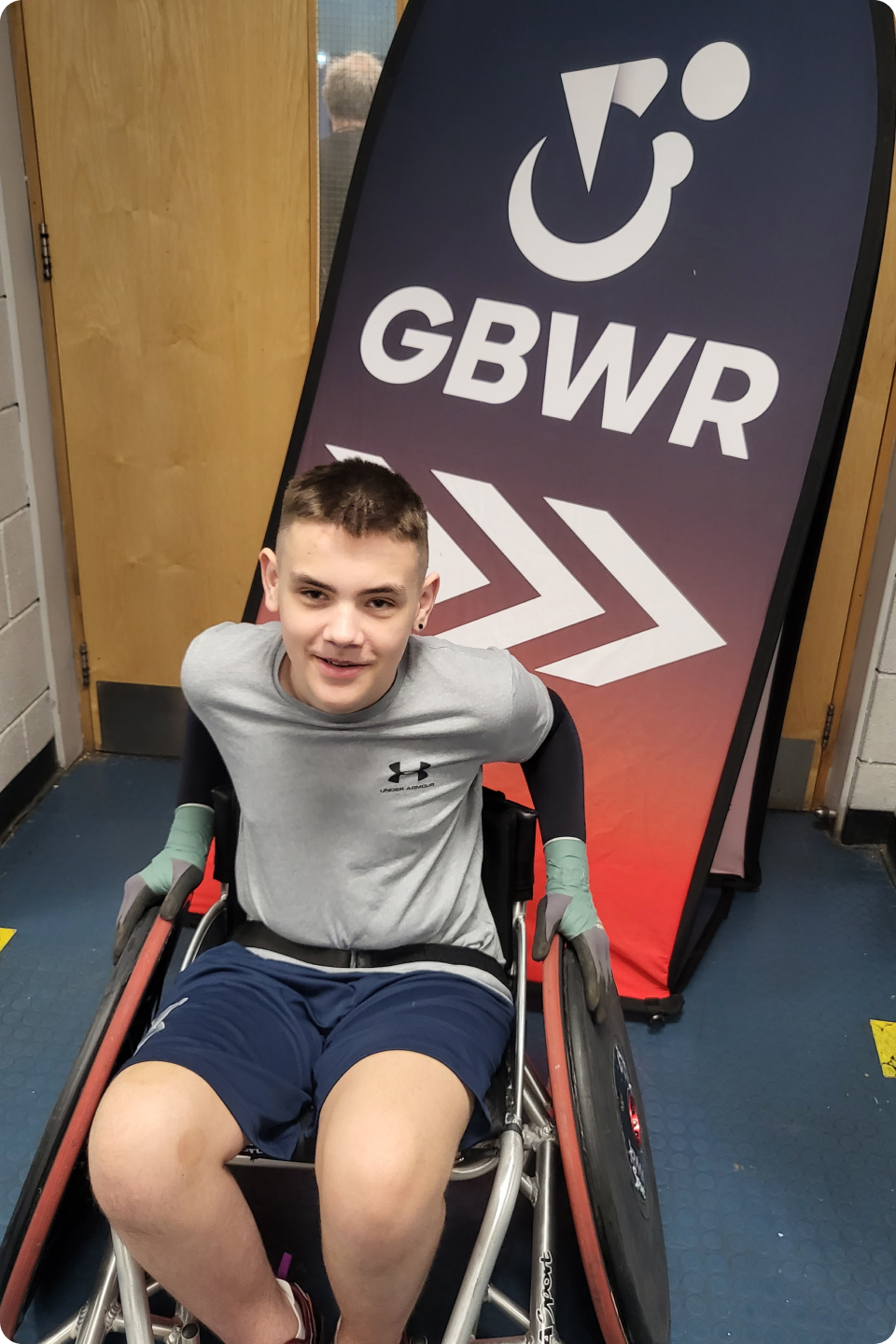 Toby Richardson in a sports wheelchair smiles at the camera. He is wearing a gray Under Armour shirt, blue shorts, and green gloves. Behind him is a banner with a wheelchair-related logo and the initials "GBWR"