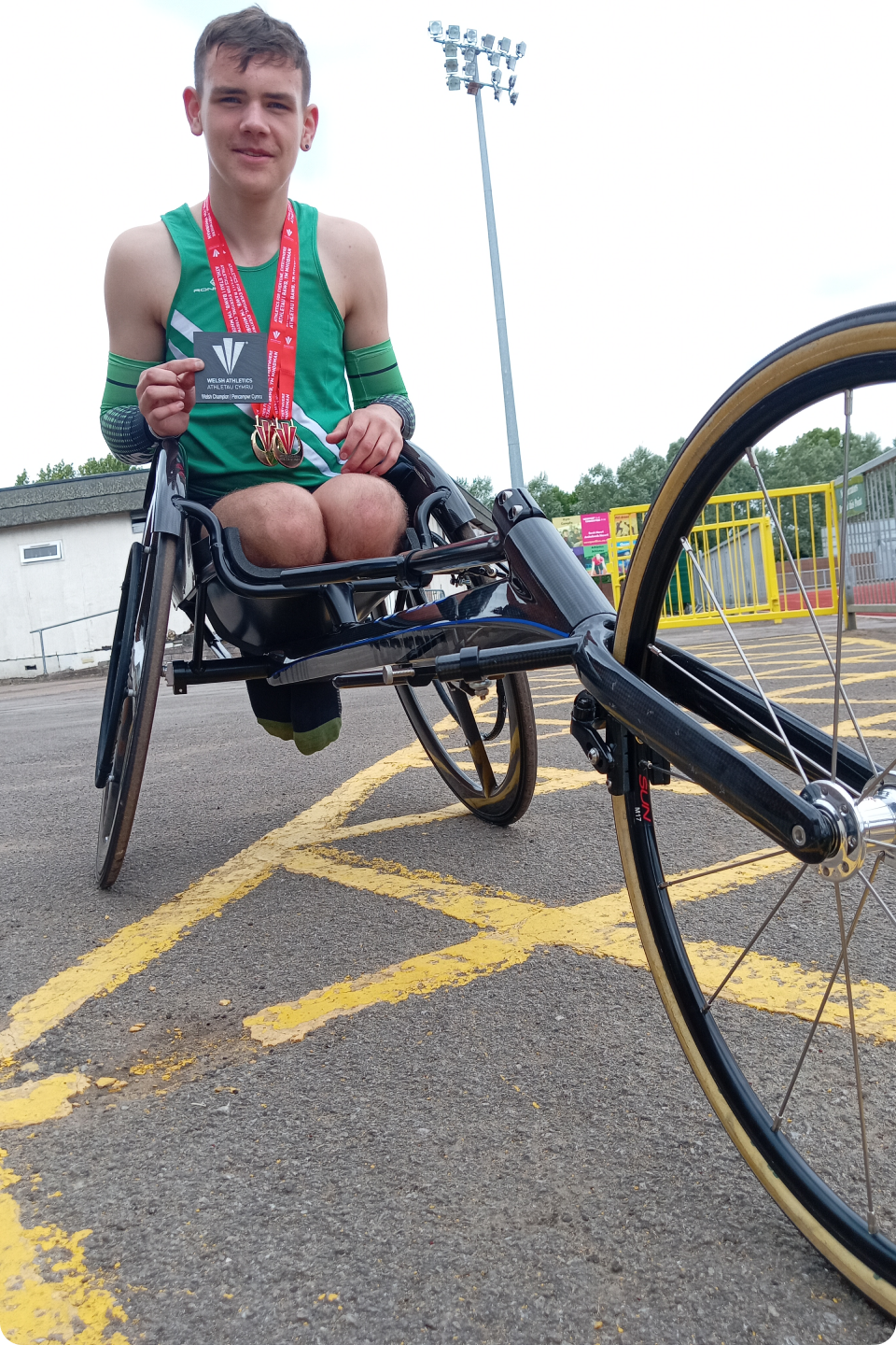 Toby Richardson in a green sports outfit is sitting in a racing wheelchair on a track, proudly displaying multiple medals around his neck. He is holding one medal up with a smile. A stadium and light posts are visible in the background.