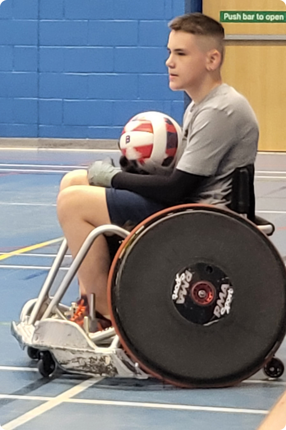 Toby Richardson in a sports wheelchair holds a red, white, and blue ball on an indoor court. They wear a gray shirt, shorts, and gloves, looking ahead with focus. The background features blue walls, and a door with a green "Push bar to open" sign.