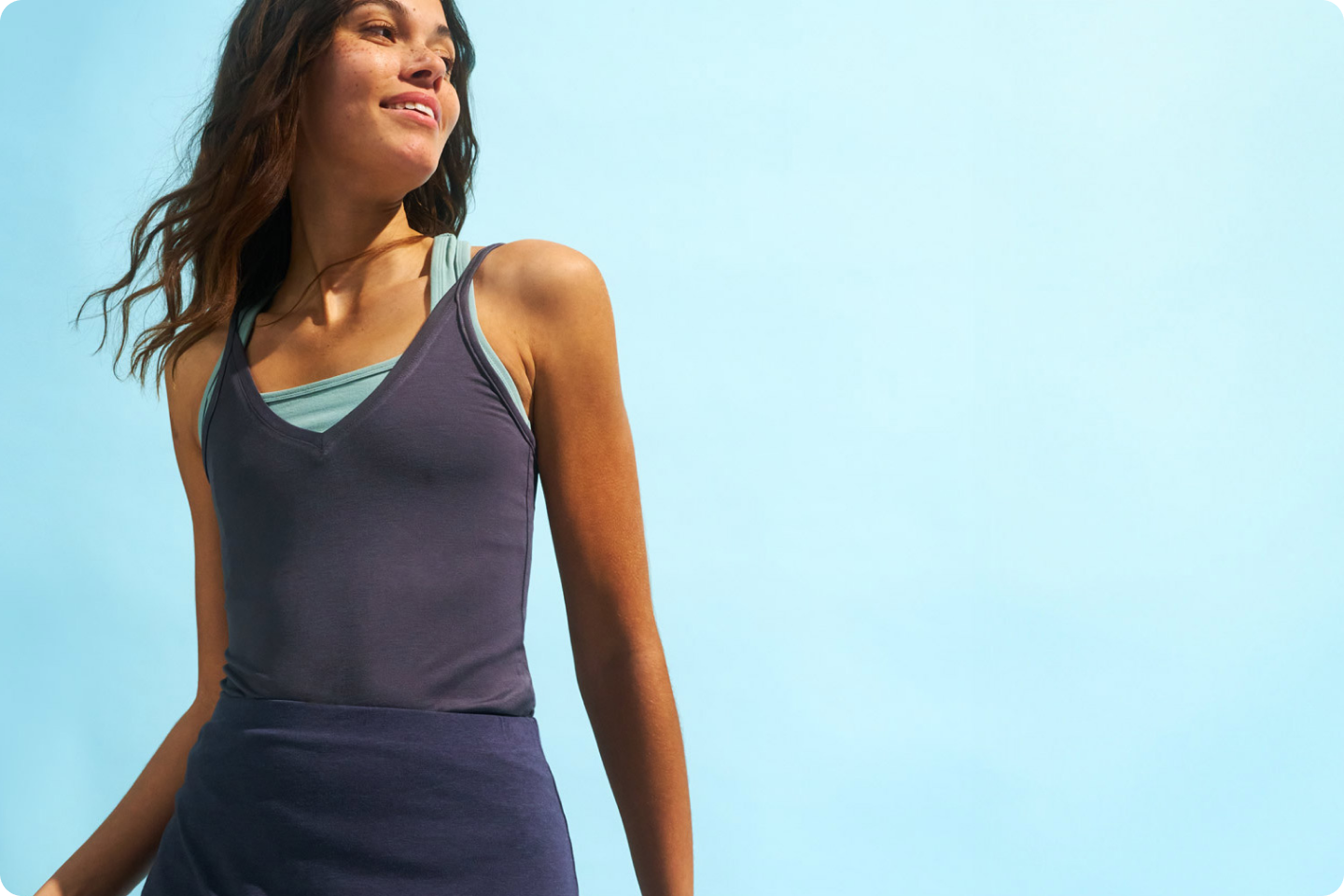 A woman with wavy brown hair is smiling and looking off to the side. She is wearing a dark blue tank top and a light blue sports bra underneath. The background is a clear blue sky.