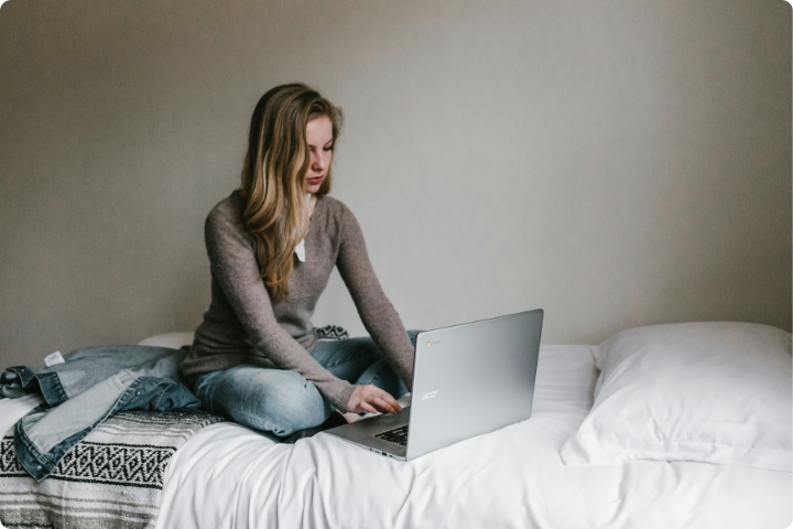 A person with long hair sits cross-legged on a bed, using a laptop. They are wearing a brown sweater and jeans. The room has a plain, light-colored wall, and there is a folded blanket and a pillow on the bed next to them.