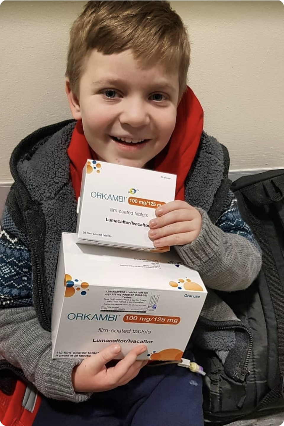 A smiling child with a light brown hair, wearing a red hoodie and patterned sweater, holds two boxes of Orkambi tablets, a medication with Lumacaftor/Ivacaftor for the treatment of cystic fibrosis. The child appears to be sitting down.