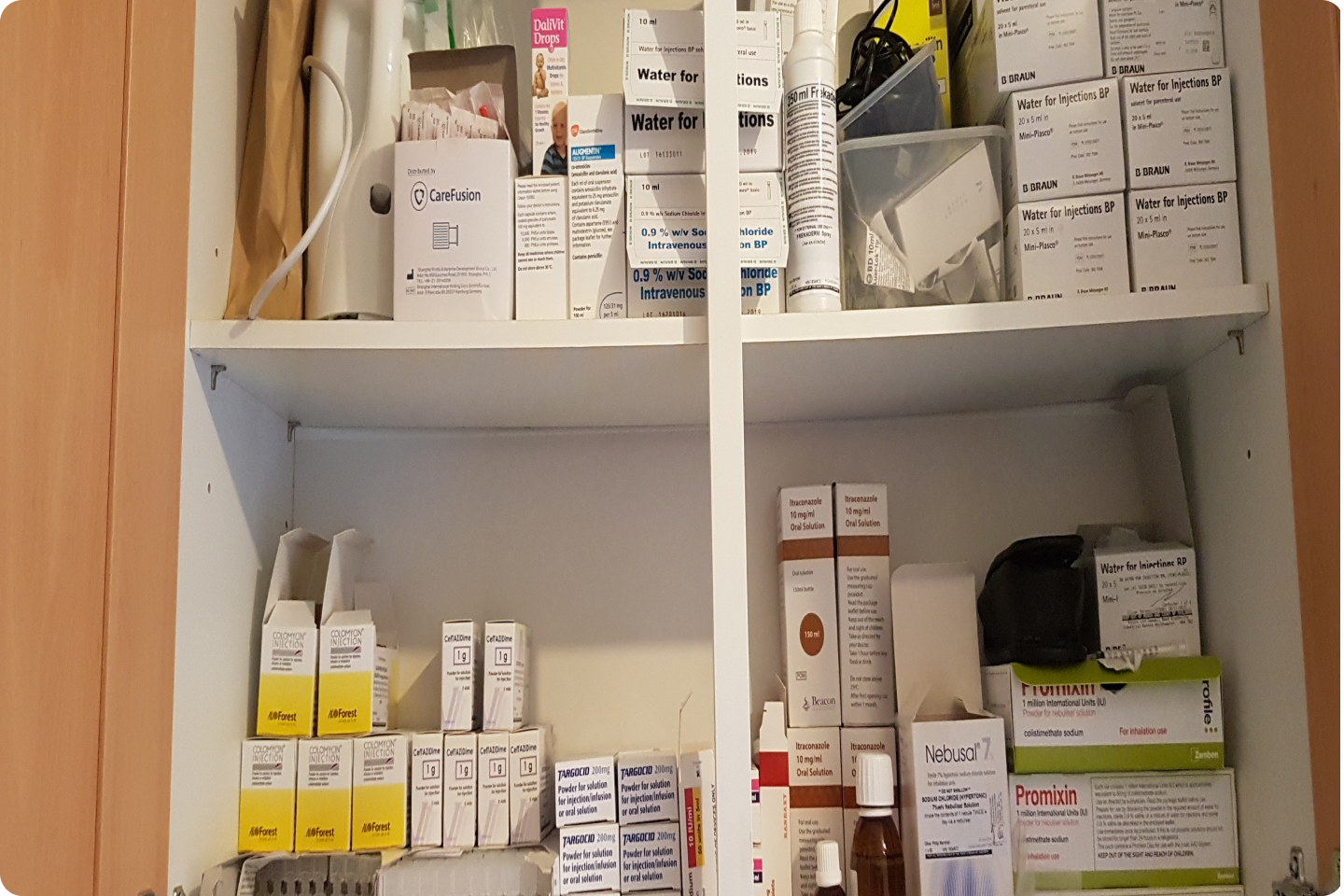 A cupboard filled with various medical supplies and medications. The shelves have boxes and bottles of different sizes and brands, including items like water for injection and Graniseptin. The supplies are organised but densely packed.