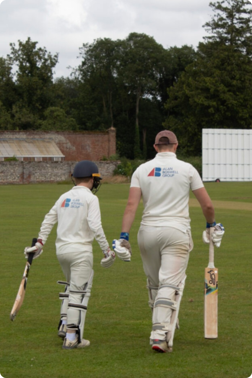 Two cricket players, one adult and one child, walk towards a field. Both dressed in white cricket uniforms and helmets, they are seen from behind, carrying cricket bats. The background includes a grassy field, trees, and sporting equipment.