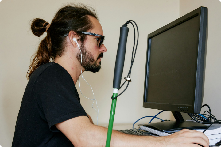 A man with long hair tied back, wearing glasses and earphones, sits at a desk using a desktop computer. He has a green cane leaning against the desk.