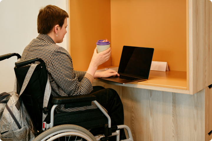 A person in a wheelchair sits at a desk working on a laptop. The person holds a reusable coffee cup in one hand and has a backpack hanging on the wheelchair. The space has a warm, orange background with wood accents.