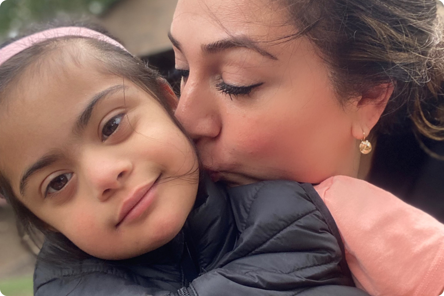 Sara hugging Amani, and kissing her on the cheek. Sara's eyes are closed and Amani is smiling. Amani is wearing a peach t-shirt and black gilet.