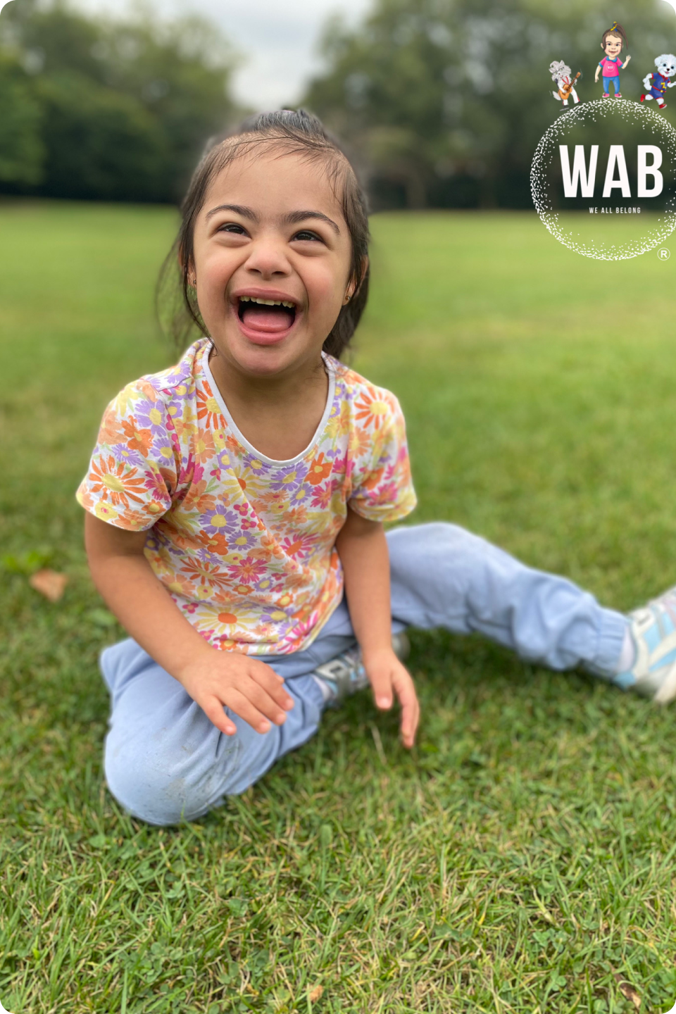 Amani sitting on grass with her left leg extended. She has a huge smile on her face. Amani is wearing a floral t-shirt and light blue joggers.