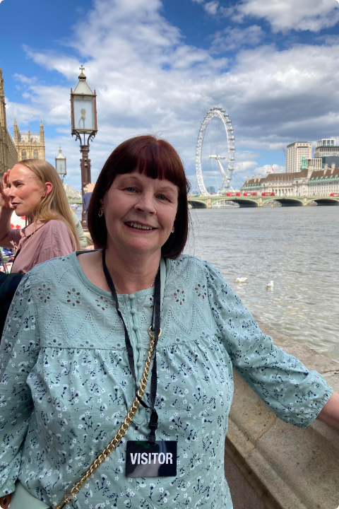 A person wearing a light blue blouse and a "visitor" badge smiles while standing by a riverside. In the background, the London Eye and historic buildings are visible under a partly cloudy sky.