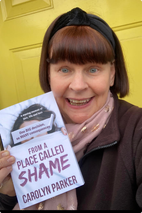 A woman with a headband and scarf holds a book titled "From a Place Called Shame" by Carolyn Parker. She is smiling, standing in front of a yellow door, and wearing a brown top. The book cover is partially visible.