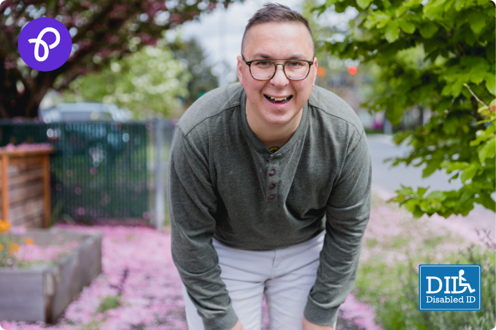 A white man is stood in a garden, he is bent forward and smiles to camera, wearing glasses, white trousers and a pale green top, in the background there are trees and cheery blossom on the ground, there are logos for Purpl discounts and disability ID DID card