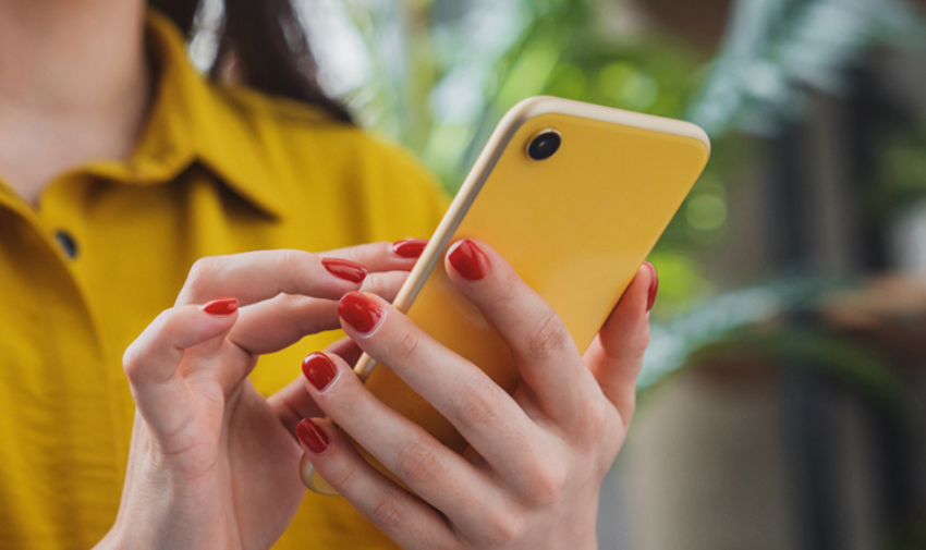 A woman wearing yellow holds a yellow phone in her hand, she is wearing red nail varnish