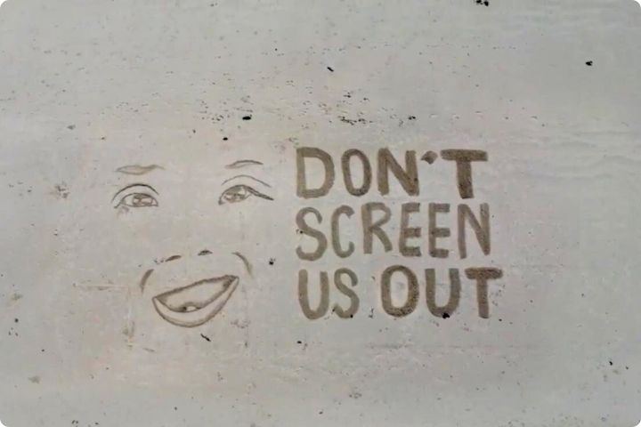 Sand drawing of a face of a person with Down syndrome and the words don't screen us out photographed from above.