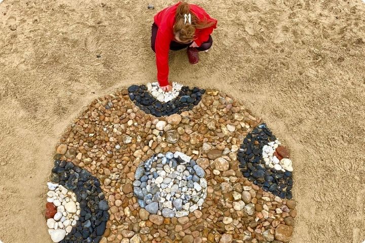 A circular piece of land art on a sandy beach made from stones and pebbles by land artist with downs syndrome Florence Halliwell who is crouched down placing a white stone and wear a red jumper.