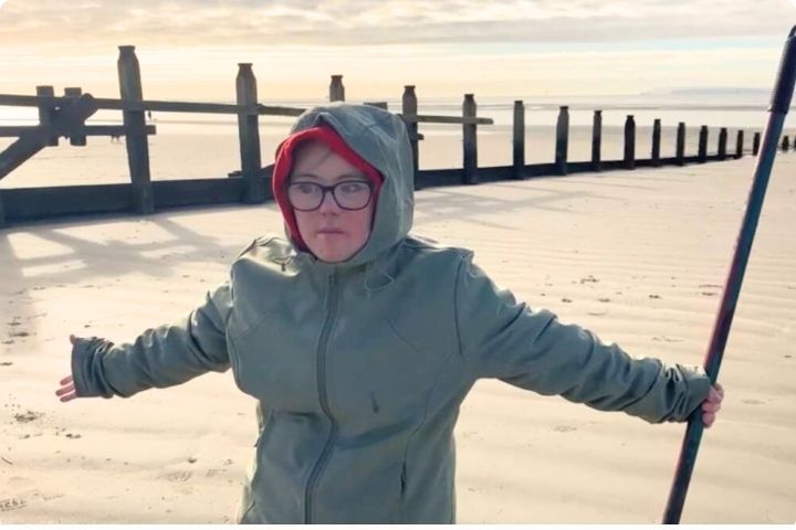 Land artist Florence Halliwell stands on a beach with arms spread, she is wearing a green hooded coat and a red hood underneath, in the background there are upright posts heading towards the sea