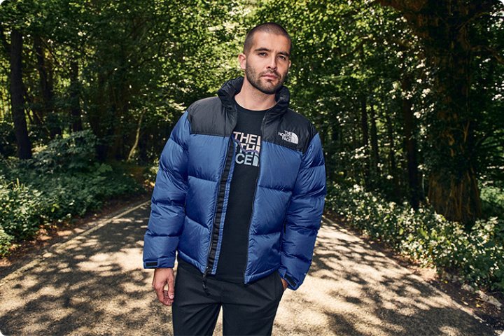 A person wearing a blue puffer jacket and a black shirt stands on a path surrounded by dense green trees in a forest. The sunlight filters through the leaves, casting dappled shadows on the ground.