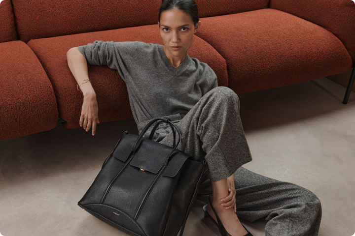 A person in a grey outfit sits on the floor, leaning against an orange sofa. They have a neutral expression and are holding a large black handbag placed beside them. The setting appears modern and minimalist