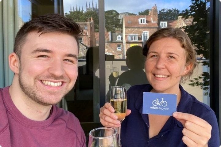 Finn on the left in a red jumper and Leah on the right in a dark blue shirt. Both are holding glasses of fizz and Leah is holding up a business card with the White Bicycle logo on it.