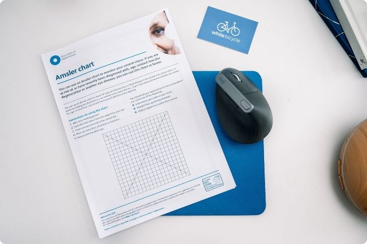 An Amsler chart - a grid which can be used to monitor changes in eyesight - on top of a blue mouse mat, next to a computer mouse and a White Bicycle business card.