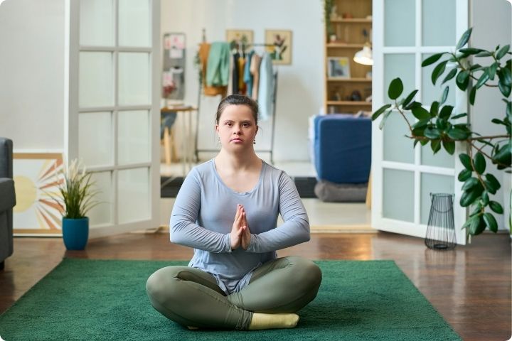 A young  woman with Down's syndrome sits in a yoga pose with legs crossed and hands in a prayer position, she is sat on a green rug in a room with plants, clothes rails and shelves in the background