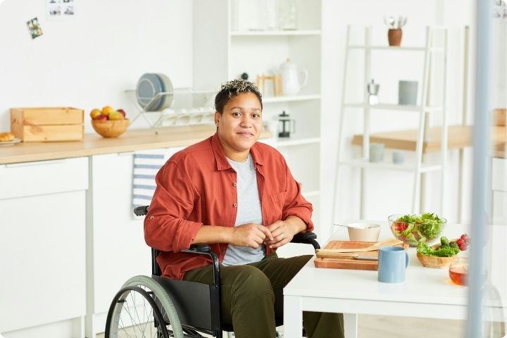 A black woman sits in a wheelchair in a kitchen, she has short cropped hair and is wearing a red shirt, grey t-shirt and dark green trousers, she is sat an a kitchen table with a salad prepared on it 