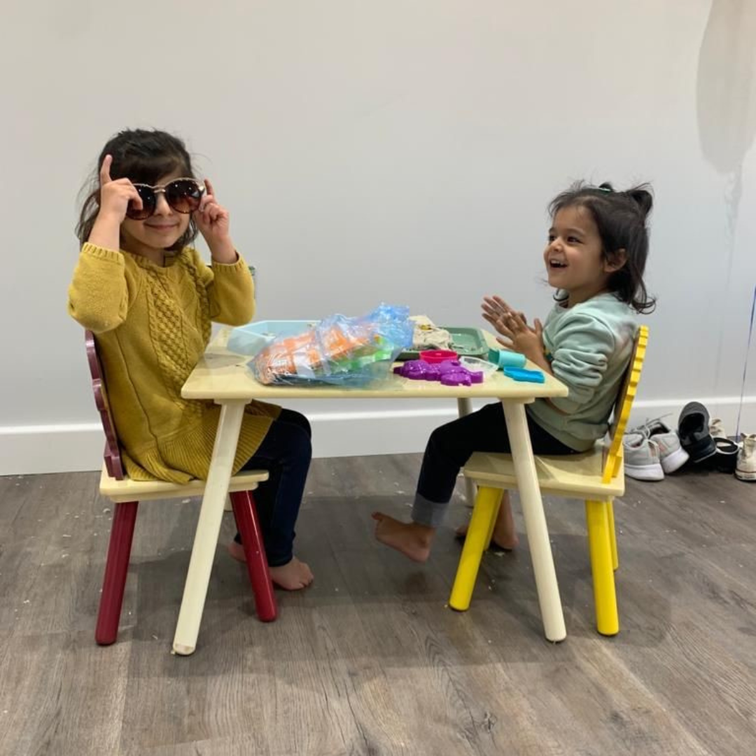 Two young girls are sitting at a small children's table, one wearing a mustard yellow sweater and the other in a pale green shirt. The girl in the yellow sweater is trying on sunglasses and looking playful, while the other girl is laughing joyfully. They appear to be engaged in a fun activity with toys scattered on the table. The setting is a bright room with a clean, wooden floor.