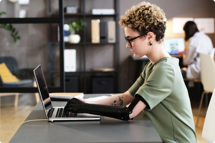 A young person who has short curly hair, glasses, a sage green t-shirt and a limb difference using a prosthetic arm sits at a desk using a laptop