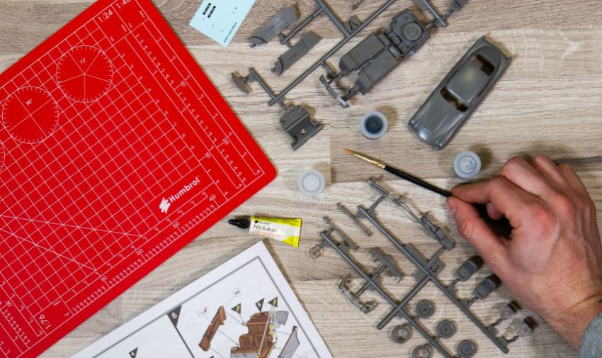 An overhead shot of an airfix car being put together, there is a hand holding a paintbrush. a bottle of glue, a red cutting mat and lots of small plastic parts