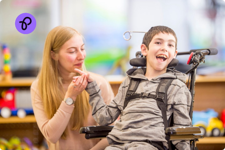 A young boy is a wheelchair user, he is wearing a harness and has brown hair and a grey top, he is smiling and holding hands with a blonde woman in a classroom