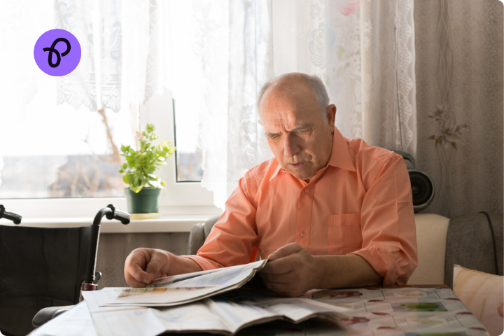 A older man has grey hair and a salmon coloured shirt, he sits at a table in his home looking at paperwork and looks stressed and confused. To the side is a wheelchair