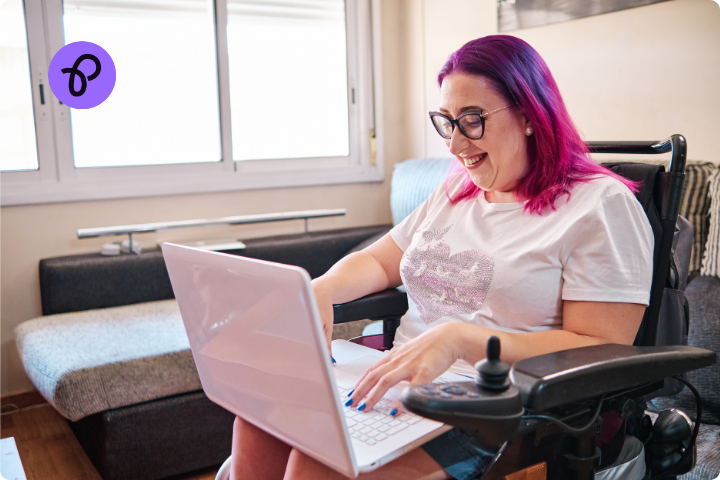 A woman with pink and purple hair is working on a laptop, she wears glasses and is a wheelchair user, she is smiling and is in her home